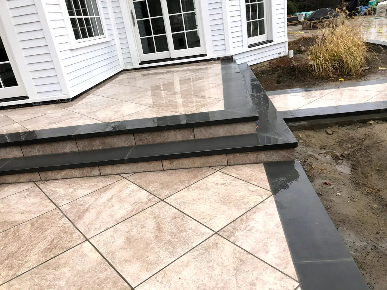 Exterior porcelain tile patio with honed bluestone step treads and risers leading up to a white shingle home with French doors, freshly rinsed and glistening