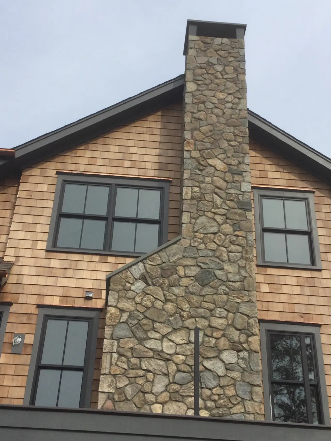 Full-height natural fieldstone chimney rising past two stories of cedar shingle siding with a metal cap