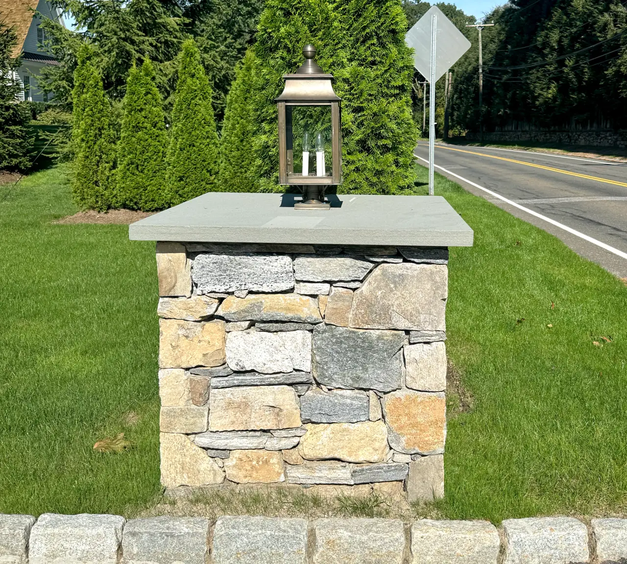 Natural stone driveway entry pillar with mixed fieldstone face, thick bluestone cap, and bronze gas lantern alongside a Belgian block curb